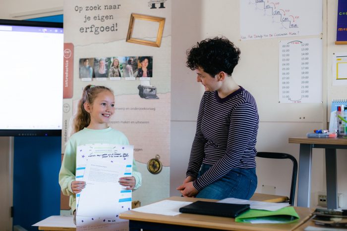 Foto van Anke Spieringhs naast een kind in een klaslokaal. Het kind lacht en houdt presentatiemateriaal vast. 