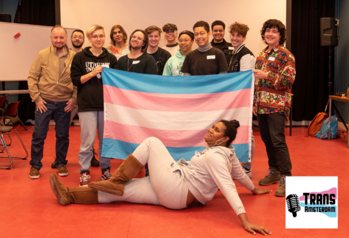Foto van mensen bij Trans Art School Amsterdam. In het midden houden ze een transgender Pride vlag vast. Rechtsonder staat het logo. 