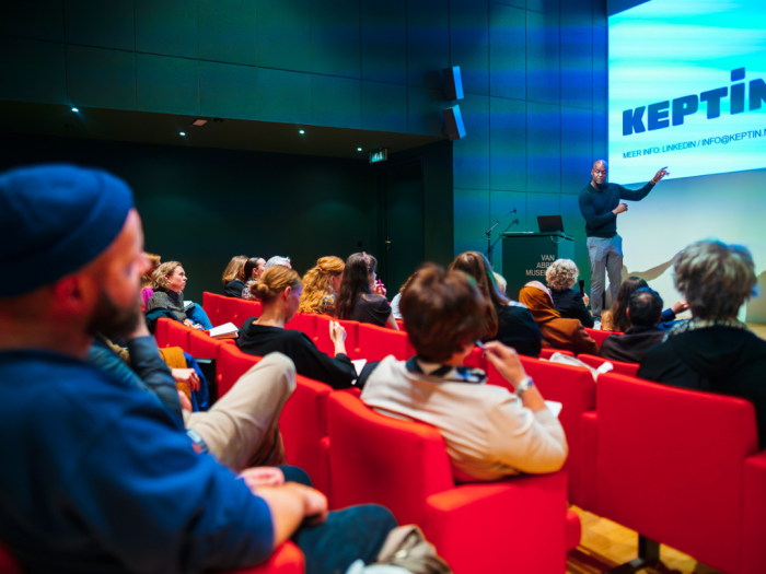 Martin van Engel in theaterzaal, spreekt voor publiek. Hij wijst naar de projectie waar 'Keptin' op te lezen is.